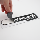 Hand pulling back a small black and white caymas boats logo decal from the background.